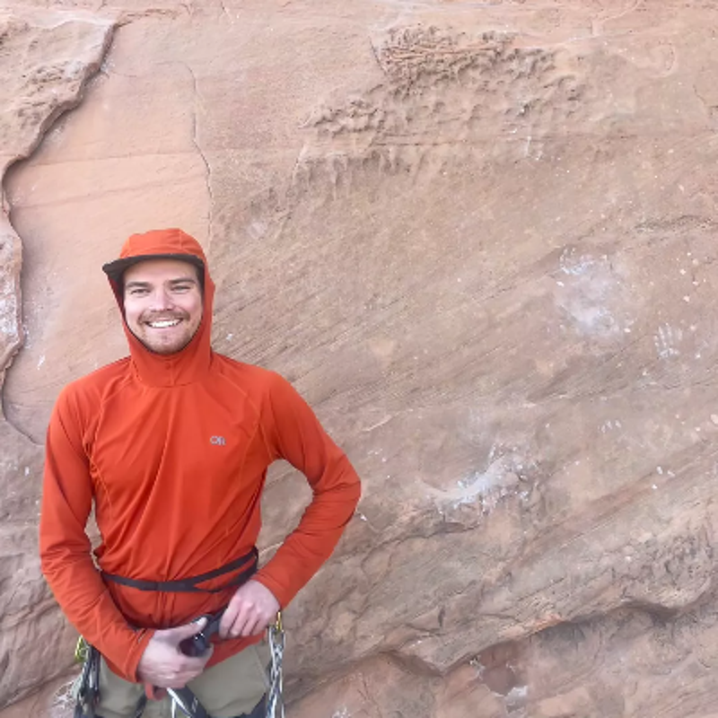 The image shows a man in an orange hooded jacket and cap, smiling at the camera. He is standing in front of a textured rock face, and he appears to be wearing climbing gear. The lighting suggests it's daytime, and the overall impression is of someone enjoying an outdoor activity.